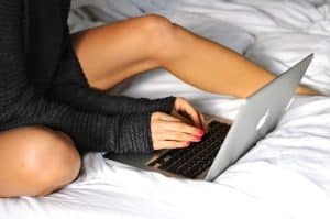 Apple Girl is using a laptop while sitting on a bed and is dressed in all-black.