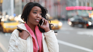 A speaking woman is standing by the side of the road wearing a white outfit and she looks lovely.