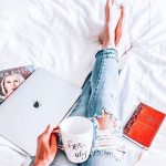 A book, coffee, and a lap are on the sexy girl's lap as she reclines in bed.