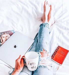 A book, coffee, and a lap are on the sexy girl's lap as she reclines in bed.