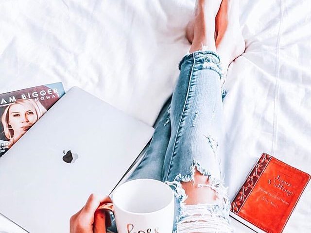 A book, coffee, and a lap are on the sexy girl's lap as she reclines in bed.